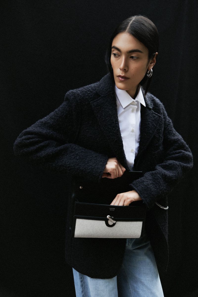 Fashion portrait of an adult woman wearing a white button-down shirt, blue jeans, and a dark jacket, reaching into a black and white bag.