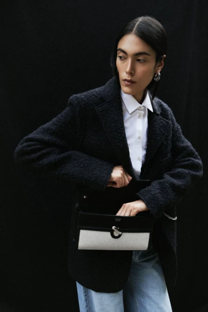 Fashion portrait of an adult woman wearing a white button-down shirt, blue jeans, and a dark jacket, reaching into a black and white bag.