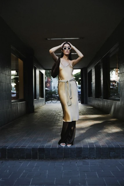 A full-length portrait of a woman in a stylish champagne-colored slip dress, sunglasses, and sandals poses in an urban tunnel.