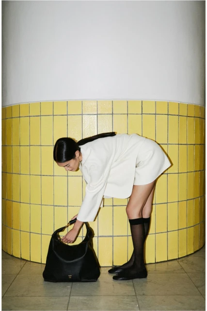 A fashionable Asian woman in a cream outfit bends down to reach into a large black handbag, next to a tiled yellow wall.