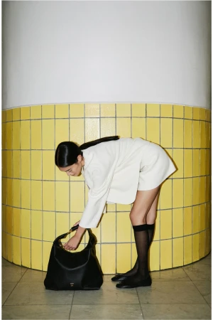 A fashionable Asian woman in a cream outfit bends down to reach into a large black handbag, next to a tiled yellow wall.