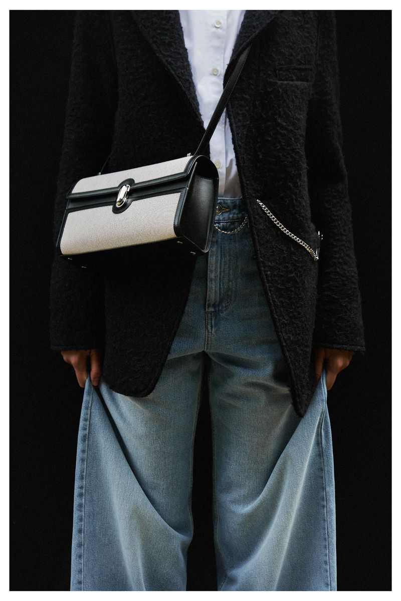 Fashion shot features handbag with black blazer, white top, and light blue jeans against dark backdrop, highlighting handbag design.