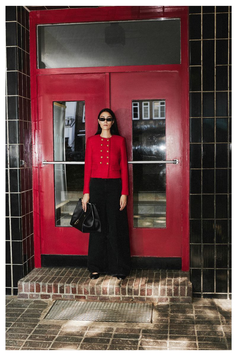 An adult woman stands in front of a red door wearing a red jacket, black pants, sunglasses, and holding a black bag.