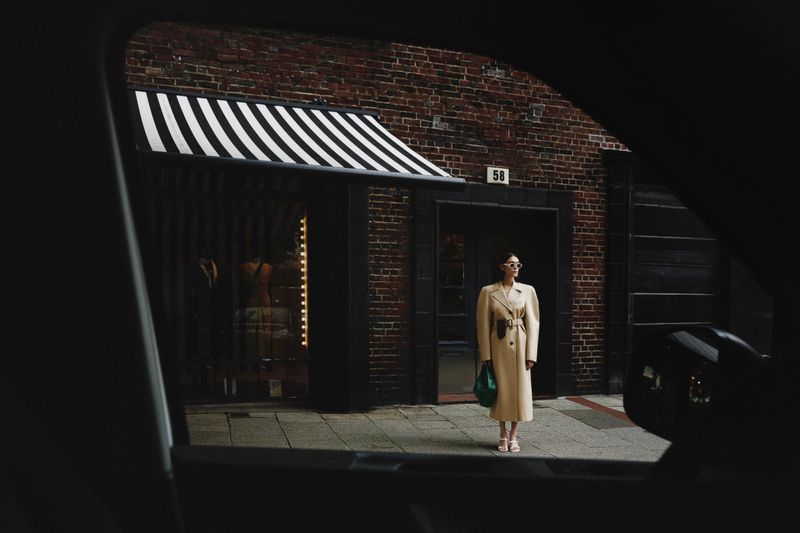 Woman in tan trench coat standing in front of a brick building with striped awning, as seen through a car window.
