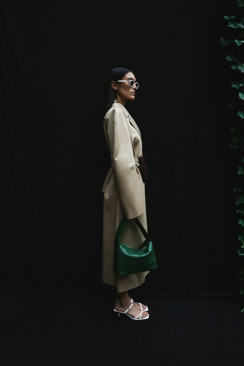 A woman models a beige trench coat, carrying a green purse, wearing white strappy heels, and sunglasses against a dark background.