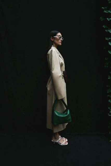 A woman models a beige trench coat, carrying a green purse, wearing white strappy heels, and sunglasses against a dark background.