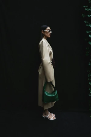 A woman models a beige trench coat, carrying a green purse, wearing white strappy heels, and sunglasses against a dark background.
