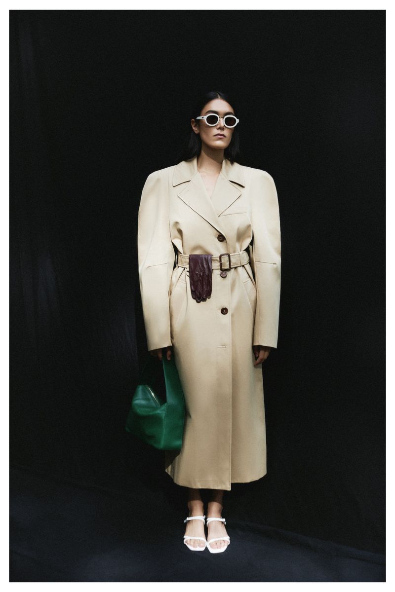 Stylish woman in a belted trench coat and sunglasses stands against a black background, accessorized with a green bag and white sandals.