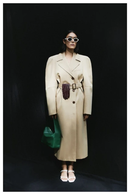 Stylish woman in a belted trench coat and sunglasses stands against a black background, accessorized with a green bag and white sandals.