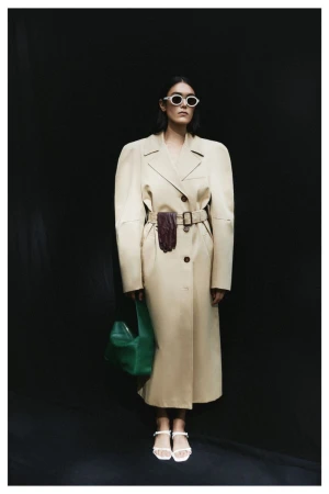 Stylish woman in a belted trench coat and sunglasses stands against a black background, accessorized with a green bag and white sandals.
