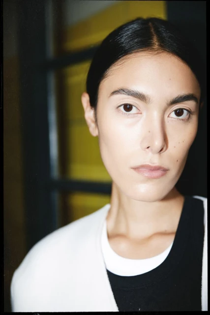 Close-up portrait of an adult woman with dark hair and fair skin wearing a white cardigan in front of a yellow interior wall.