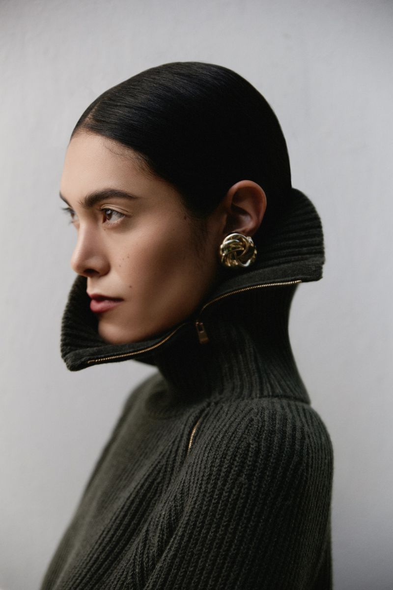 Elegant fashion portrait of a woman with dark hair wearing a zipped dark green turtleneck sweater, gold earrings, posing in a studio.