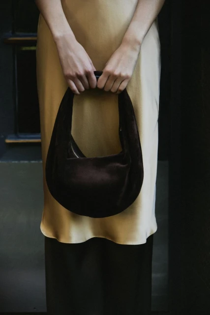 A woman in a beige slip dress holds a dark brown velvet handbag, presenting a minimalist fashion and texture study.