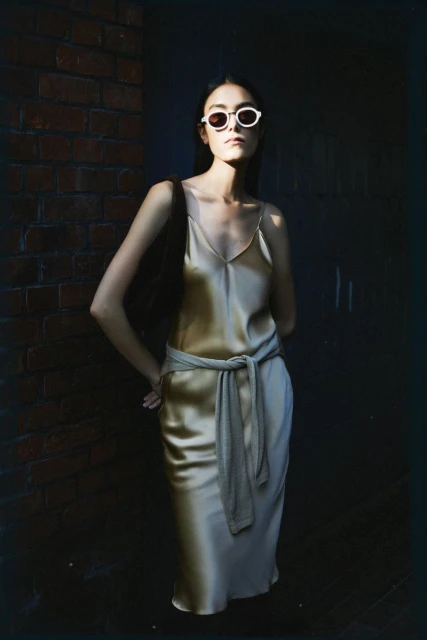 A slender woman with sunglasses poses in a champagne silk dress with a tied waist, against a textured brick and dark wall, bathed in natural light.