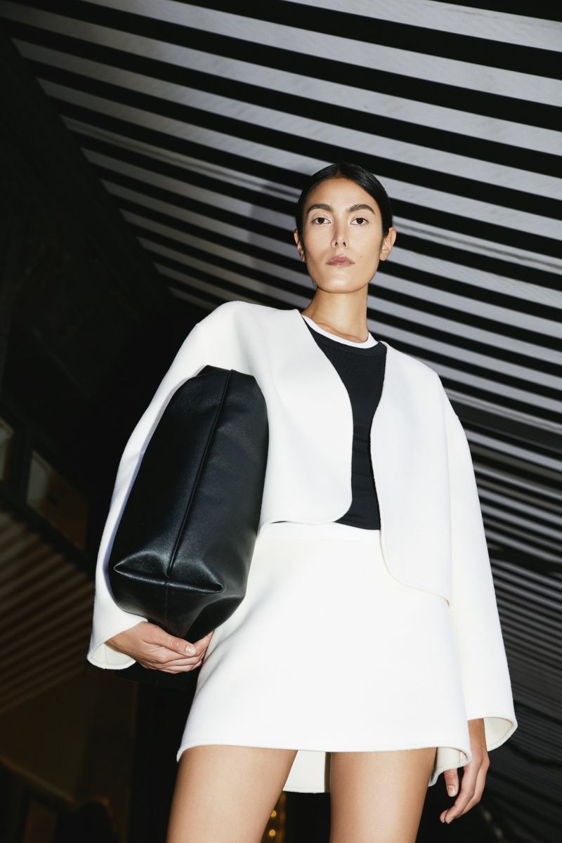 Fashionable woman in a white skirt suit poses with a black tote bag against a striped ceiling, creating a high-fashion visual.