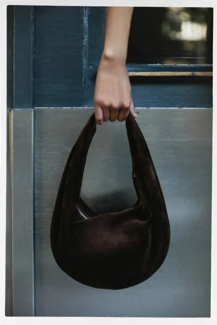 Close-up studio shot of a dark brown suede hobo bag held by a fair-skinned hand, against a blue and grey metal background.