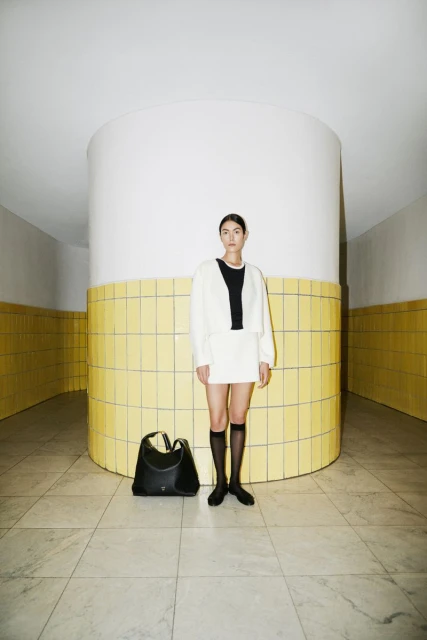 Woman in cream jacket and skirt poses with a black handbag in front of a column with yellow tiles in an indoor setting.
