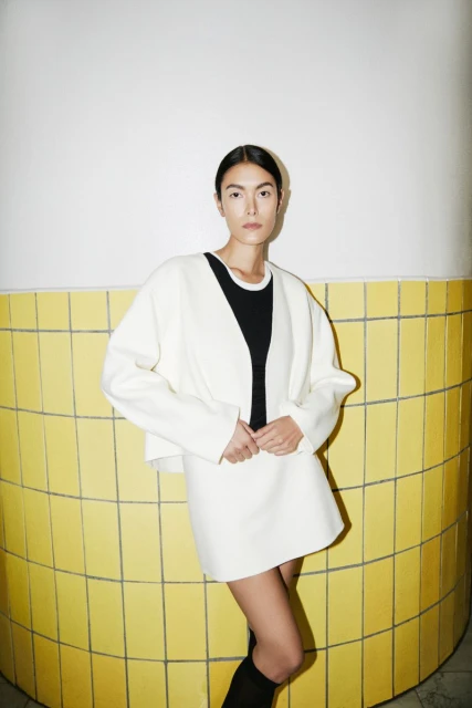 Fashionable woman in a cream-colored jacket and skirt stands against a tiled wall in a studio, showcasing a minimalist and elegant style.