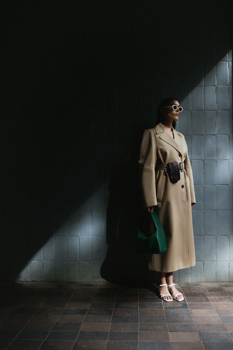 Model wearing a beige belted coat and sunglasses holding a green handbag against a tiled wall outdoors in sunlight and shadow.