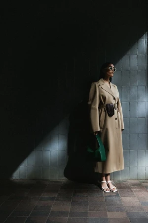 Model wearing a beige belted coat and sunglasses holding a green handbag against a tiled wall outdoors in sunlight and shadow.
