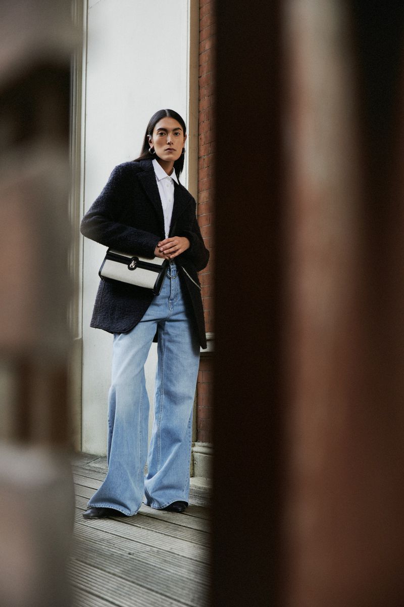 A stylish adult woman is posing in a black blazer, white shirt, and blue jeans, holding a purse with a blurred background. Fashion shot.