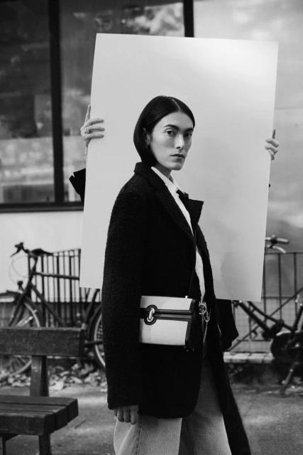 Black and white fashion shot of a woman in a black coat holding a cream-colored bag and a white display board in an urban environment.