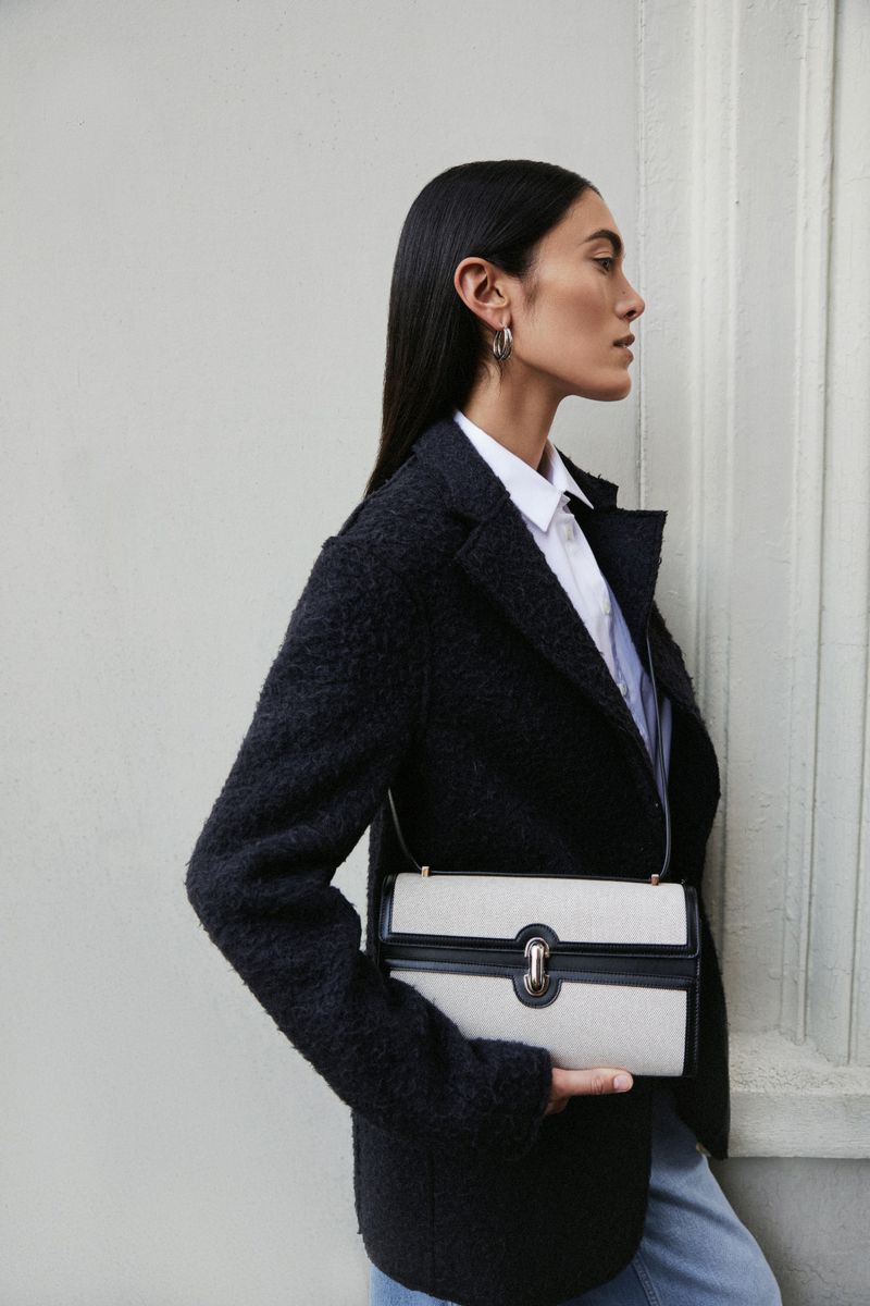 Woman in dark textured jacket and white shirt holding a light-colored handbag against a plain wall.