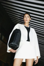 Model in white jacket and skirt holding a black handbag against a striped ceiling background for Anita Hass Fall 2023.