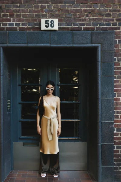 Model in a gold silk slip dress standing in front of a dark doorway beneath a brick building exterior with building number 58.