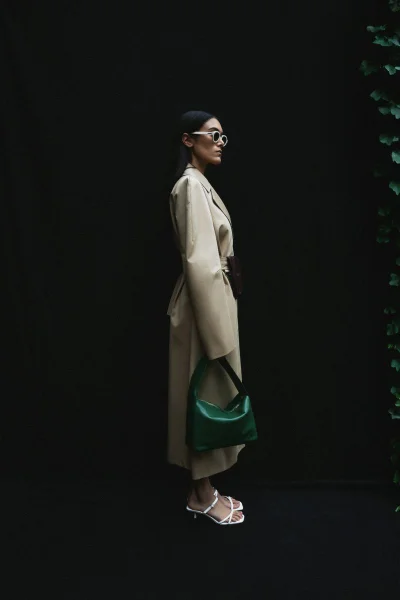 Model in profile wearing a beige trench coat, white frame sunglasses, and white sandals holding a green handbag against a black background.