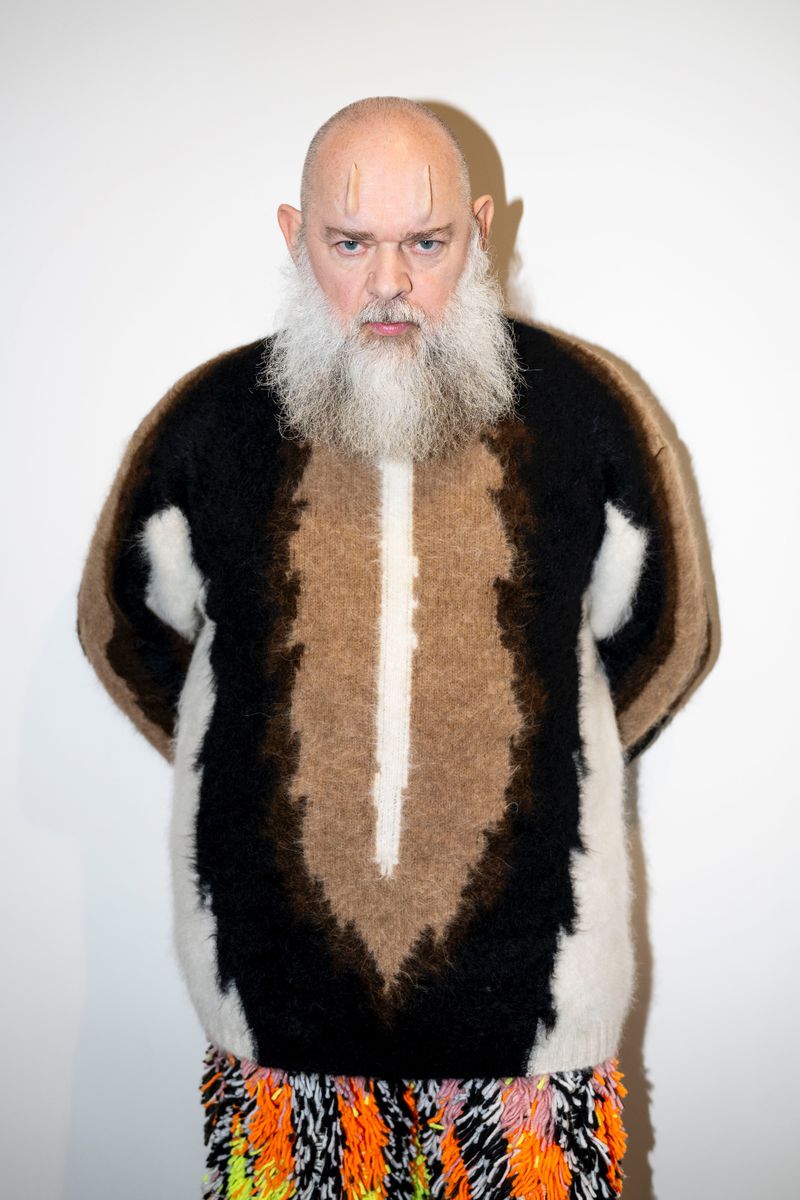 Portrait of a bald adult man with a long white beard wearing an eye-catching sweater with a black, brown, and white pattern.