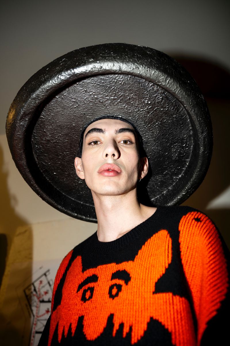 Man in an orange and black sweater poses with a textured black hat in an indoor setting, captured with a medium shot.