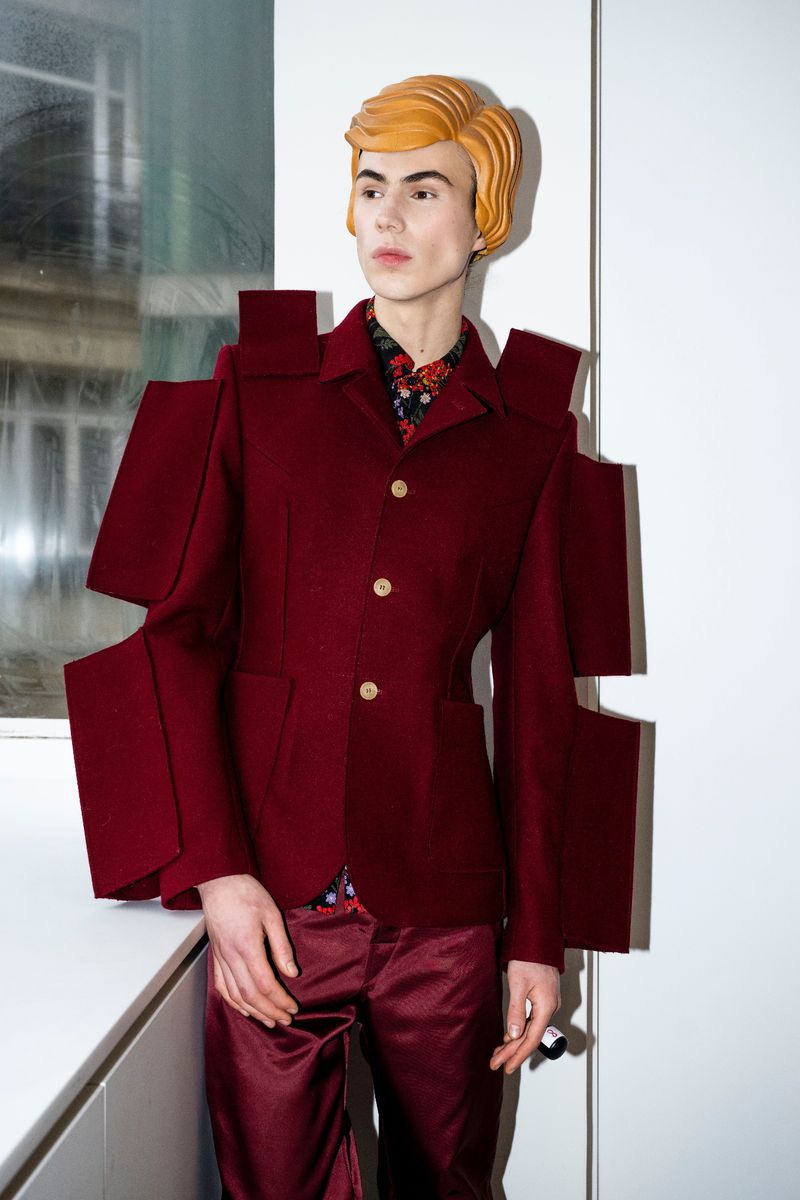 A person with fair skin and strawberry-blonde hair wears a maroon jacket and trousers in a stylish portrait in an indoor environment.