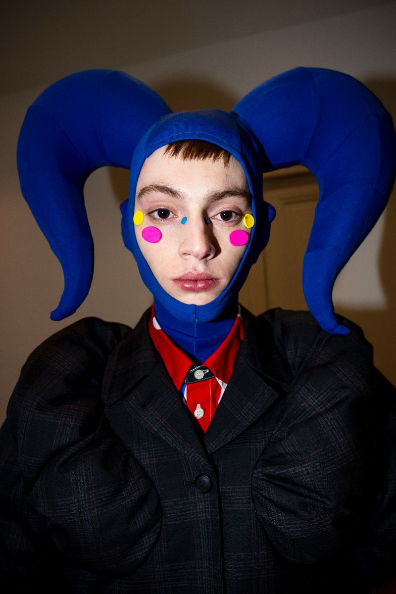A person in a blue headdress with pink and yellow face paint, wearing a red shirt and black blazer, poses indoors.
