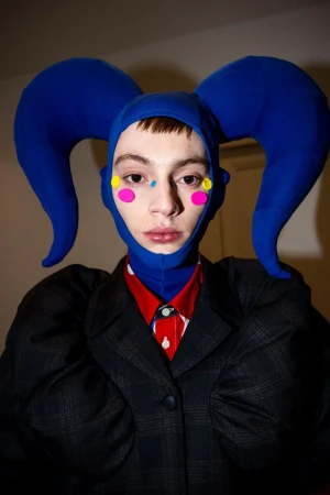 Model wearing a royal blue horned hood, face paint dots, and a dark plaid jacket backstage at Walter Van Beirendonck FW24.