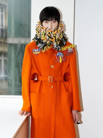 Model wearing a bright orange belted coat with patches and a multicolor textured high-neck collar backstage.