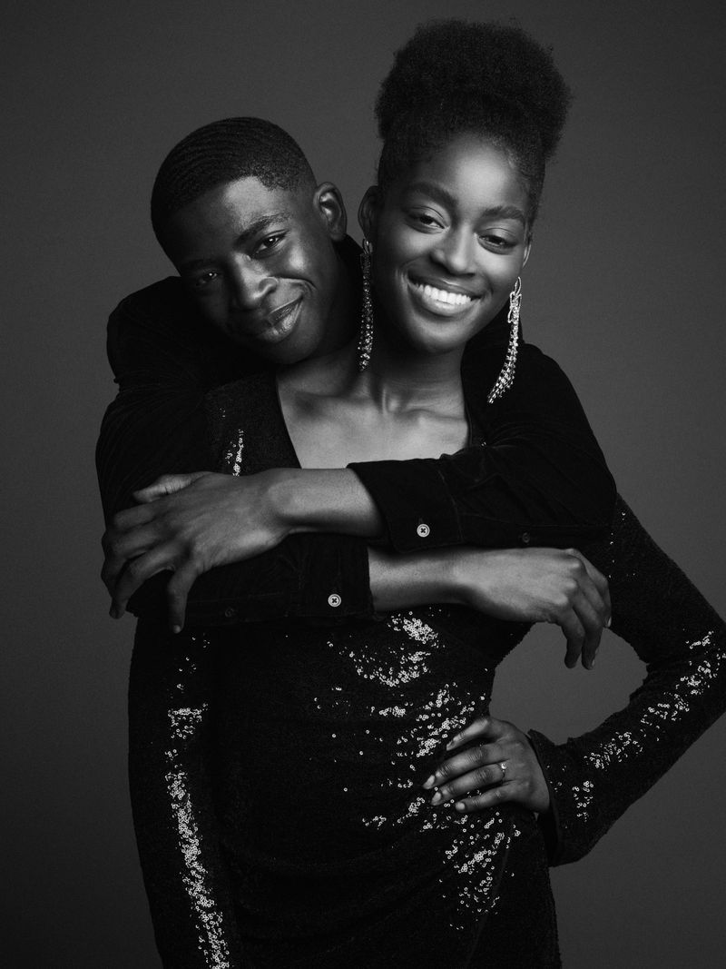 Portrait of a happy couple: an African American woman in a sequined dress smiles as an African American man embraces her in front of a gray backdrop.