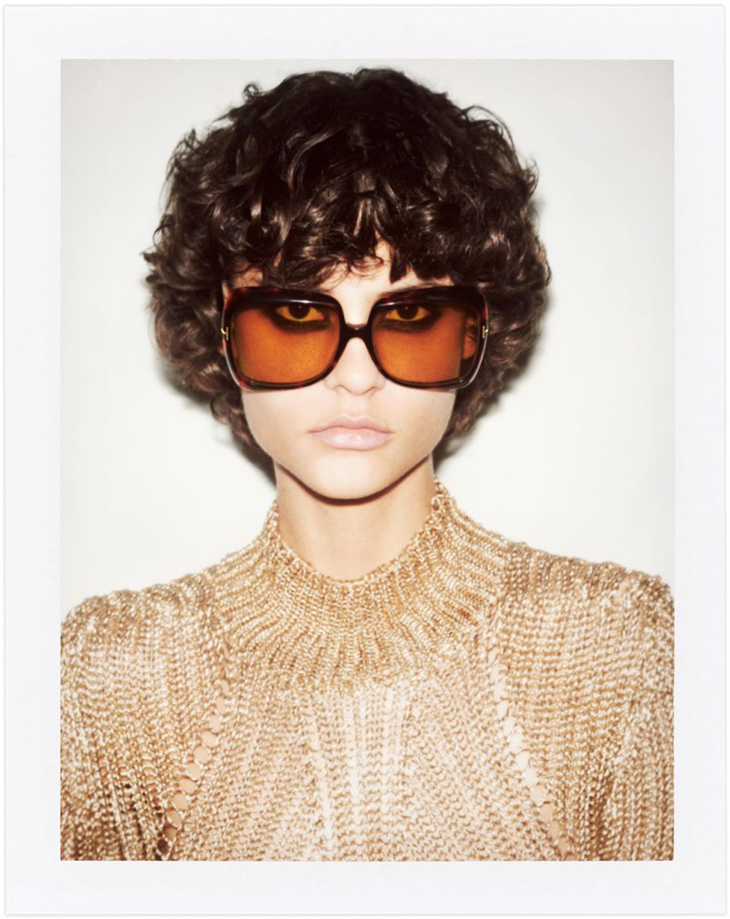 Close-up studio shot of a young female model with curly hair wearing oversized sunglasses and a metallic gold knit top with a high neckline.
