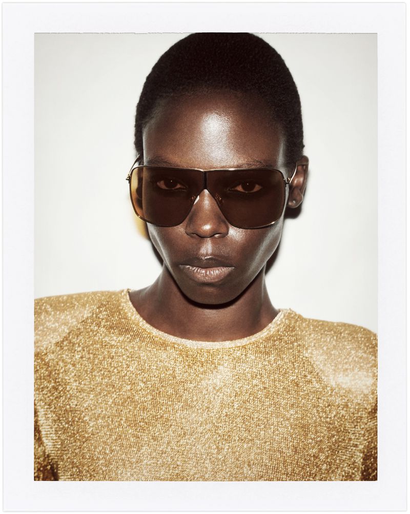 A close-up studio shot of an African woman with short hair wearing sunglasses and a shimmering gold top against a bright white background.