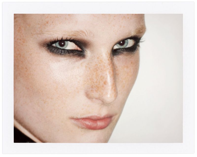 Close-up portrait of an adult female with fair skin, freckles, and blue eyes accentuated with heavy dark makeup and light eyebrows.