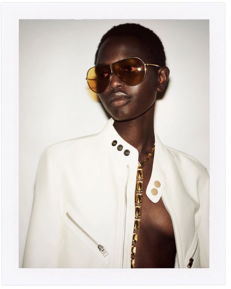 A dark-skinned model poses indoors wearing aviator sunglasses, a white jacket, and a gold necklace against a white background.