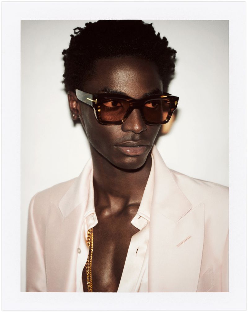 Close-up portrait of an adult man with dark skin wearing amber-tinted sunglasses, a light pink suit jacket, and a gold chain necklace.