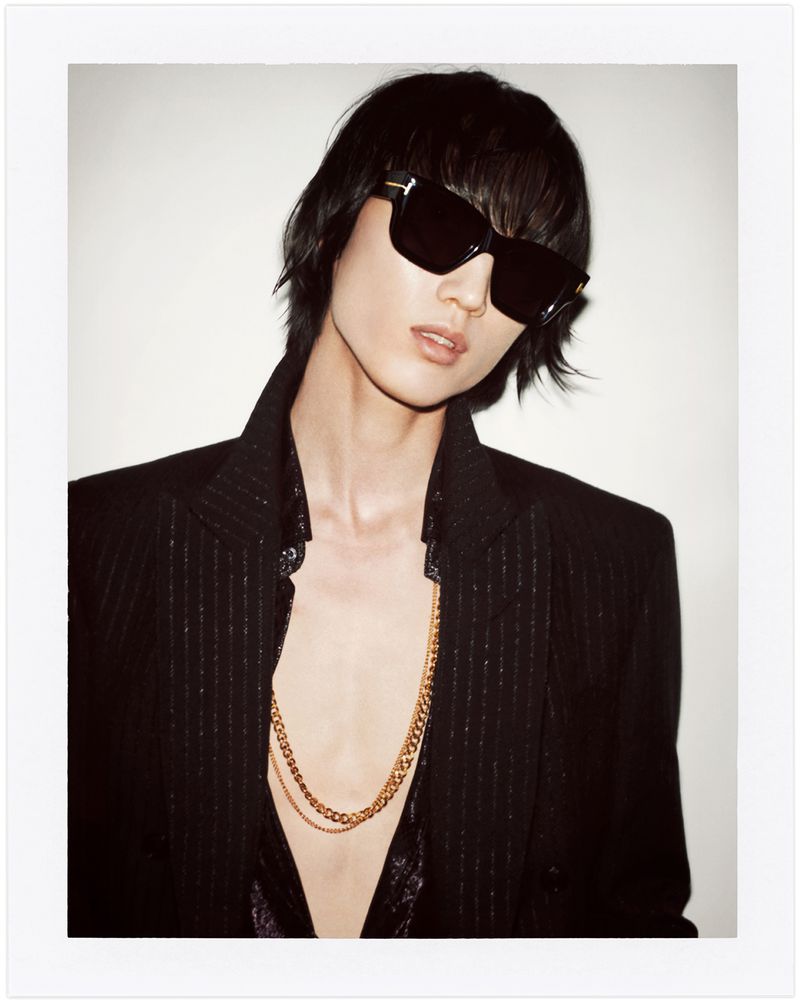 Portrait of an adult female wearing sunglasses and a dark blazer with a gold chain necklace in front of a light-colored background.