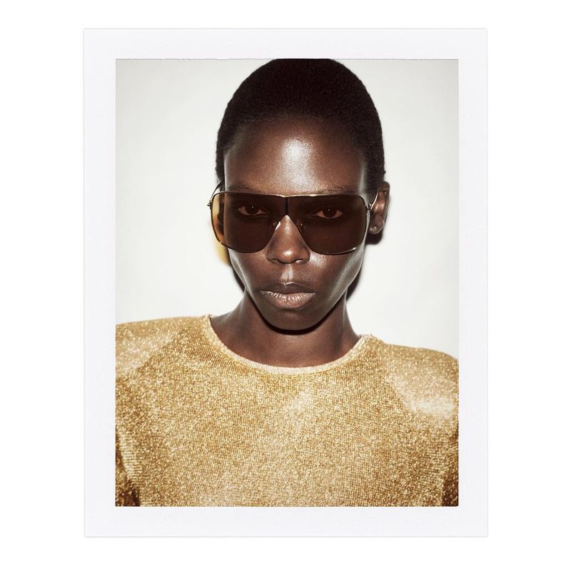 Headshot of a black female model wearing sunglasses and a gold sparkling top against a white background in a studio setting.