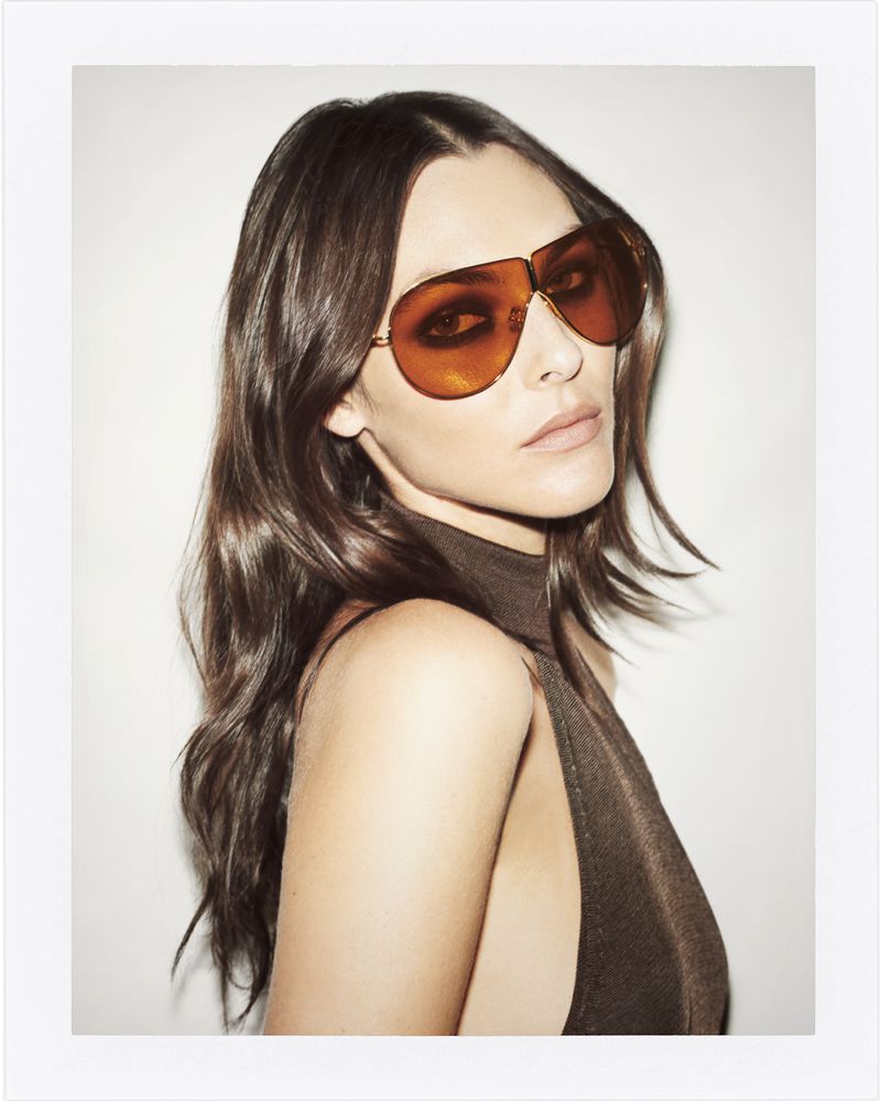 Portrait of a woman with long brown hair wearing a brown top and large aviator sunglasses against a white background in a studio.