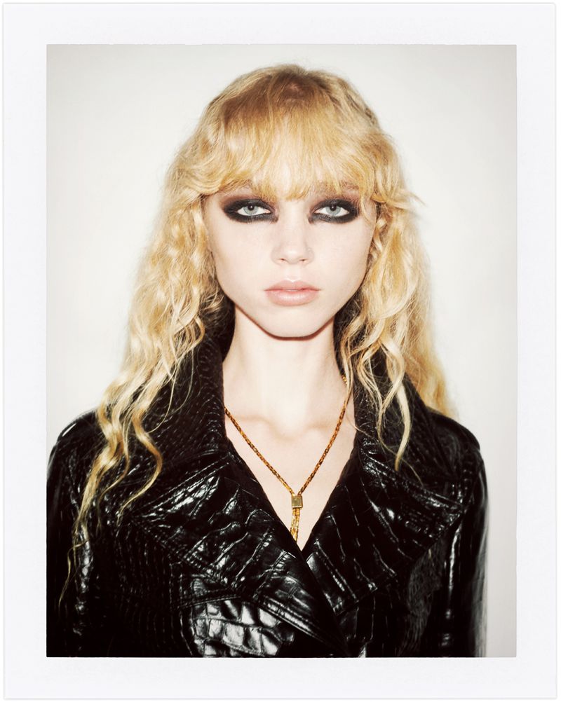 Blonde woman with heavy eye makeup wearing a black, reptile-textured coat and gold necklace against a stark white backdrop.