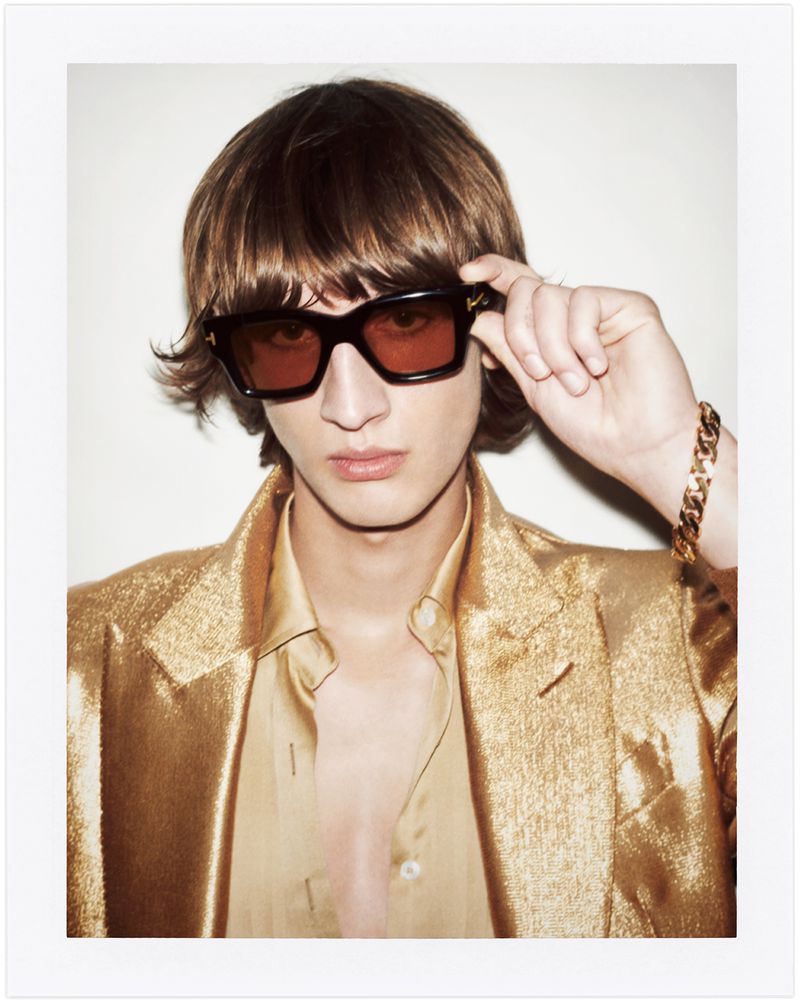 A young adult with sunglasses and a gold jacket poses indoors against a white wall. A bold gold chain and warm lighting complete the look.