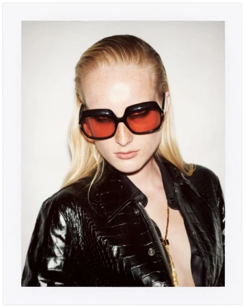 Model wearing oversized tinted sunglasses and a glossy black embossed leather jacket against a light backdrop in a polaroid frame.