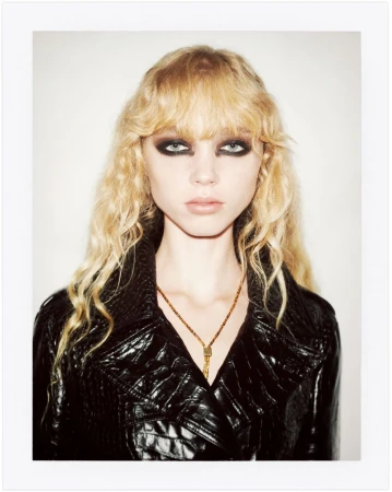 Model with heavy dark eye makeup and wavy blonde hair wearing a black patent jacket against a plain background