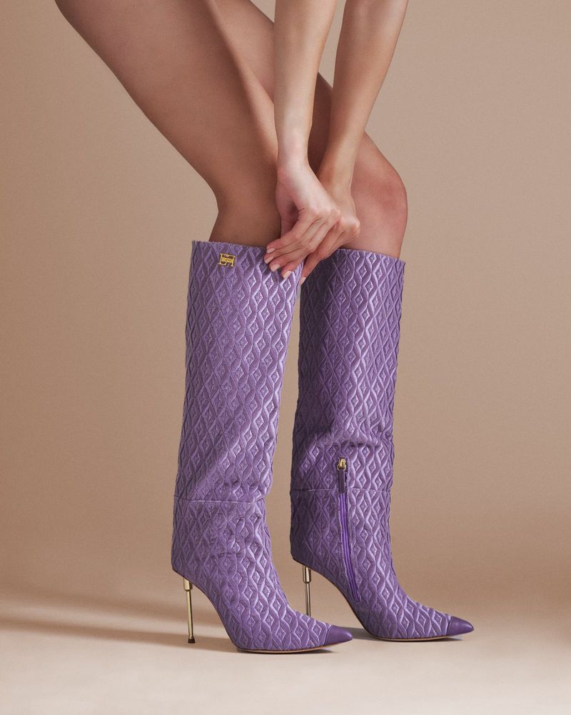 Close-up of a woman's legs and hands adjusting textured lavender heeled boots against a neutral background in a studio setting.